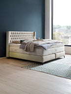 Ein luxuriöses Boxspringbett mit getuftetem Kopfteil in einem Schlafzimmer mit dunkelblauer Wand und großem Fenster.