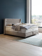 Ein luxuriöses Boxspringbett mit gepolstertem Kopfteil in einem modernen Schlafzimmer mit Holzboden und blauem Teppich.