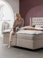 Lächelnde Frau sitzt auf einem Bett mit beigem Kopfteil, liest ein Magazin. Im Hintergrund ein Kleiderständer.