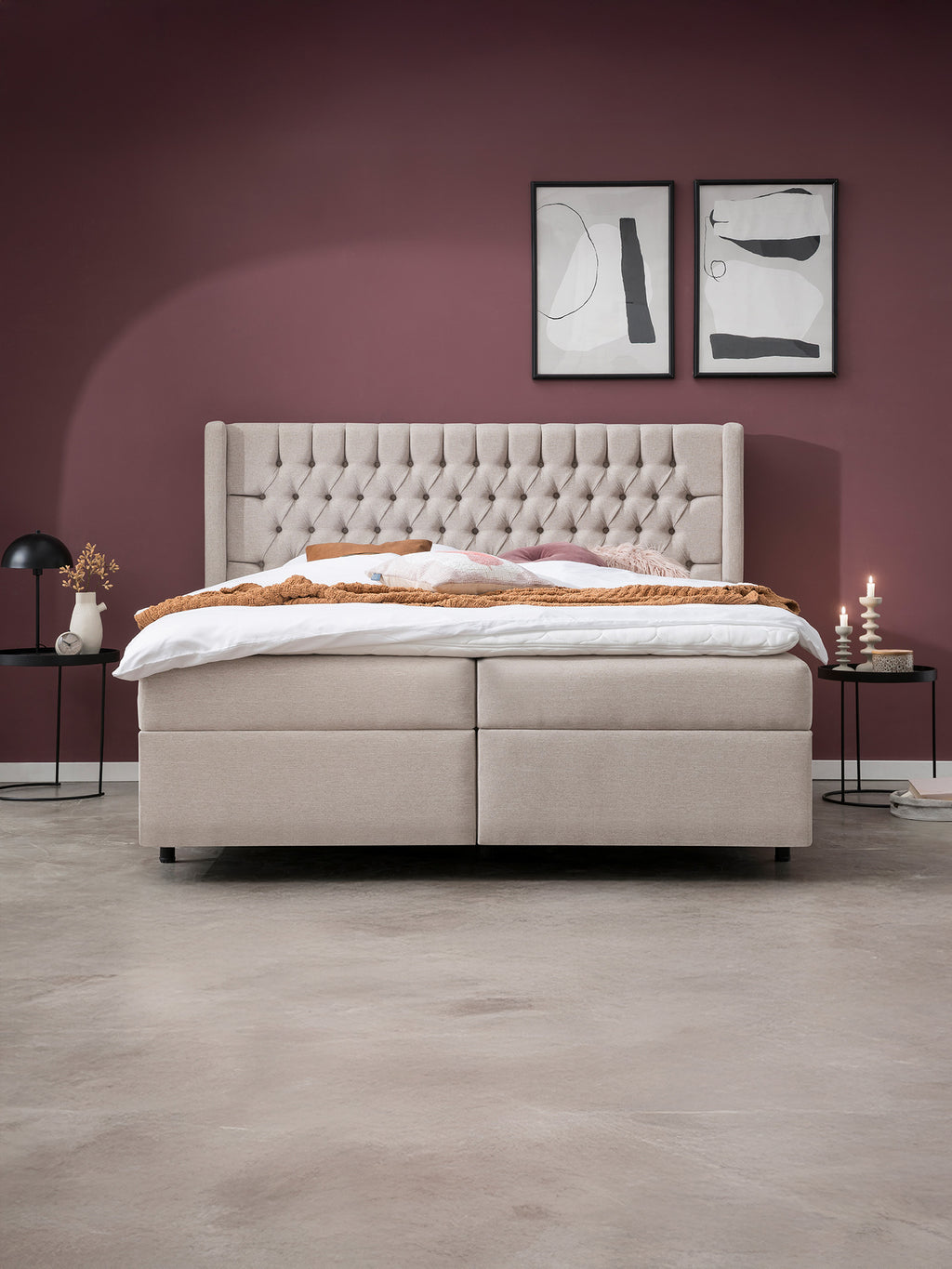Ein beiges Boxspringbett mit getuftetem Kopfteil steht vor einer weinroten Wand. Zwei abstrakte Bilder hängen darüber.