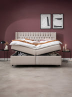 Ein beiges Boxspringbett mit Stauraum, der geöffnet ist, steht in einem Schlafzimmer mit weinroten Wänden.