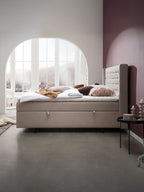 Ein Schlafzimmer mit einem Boxspringbett, das mit weißen Laken und verschiedenen Kissen in Erdtönen bezogen ist. Im Hintergrund ist ein großes Bogenfenster mit Blick auf eine Stadt und Berge zu sehen. Die Wände sind weiß und lila. Ein kleiner runder Tisch mit Kerzen steht neben dem Bett.