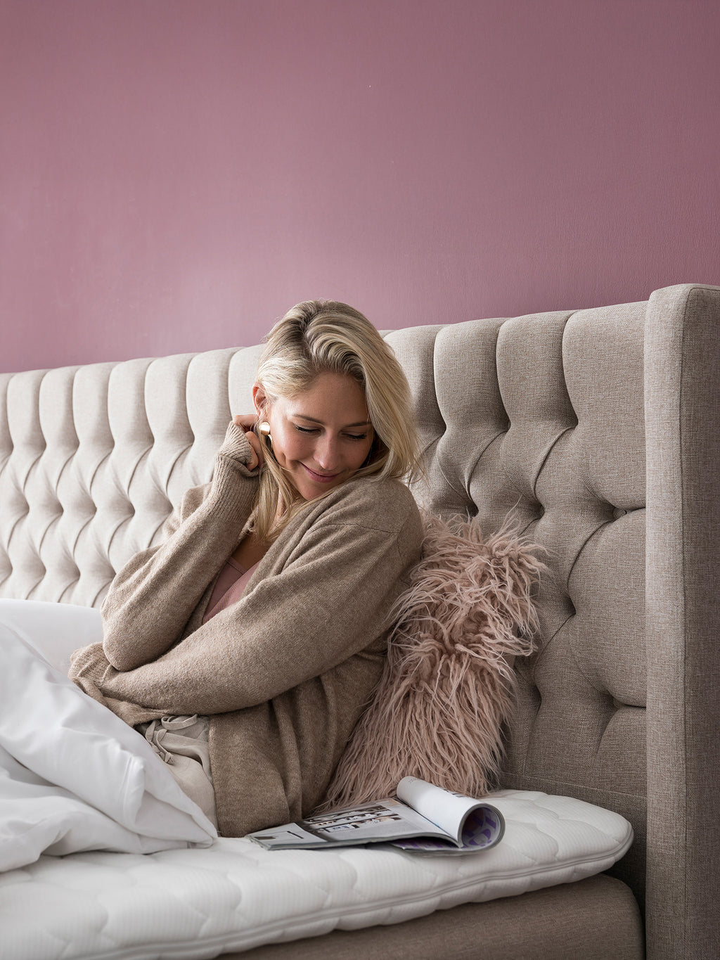 Frau sitzt lächelnd im Bett, trägt einen beigen Cardigan und schaut auf eine Zeitschrift.
