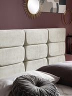 Gepolstertes Kopfteil in hellem Beige mit Kissen auf dem Bett. Eine goldene Spiegel-Sonne hängt an der Wand.
