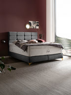 Graues Boxspringbett mit gepolstertem Kopfteil und weißer Matratze in einem Schlafzimmer mit weinroter Wand.
