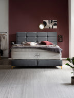 Ein luxuriöses Boxspringbett mit grauem Kopfteil und weißer Matratze in einem Schlafzimmer mit weinroter Wand.
