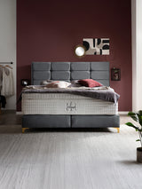 Ein luxuriöses Boxspringbett mit grauem Kopfteil und weißer Matratze in einem Schlafzimmer mit weinroter Wand.