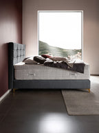 Ein luxuriöses Bett mit einem grauen Kopfteil und einer weißen Matratze, umgeben von Kissen und einer Decke.