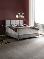 Graues Boxspringbett mit gepolstertem Kopfteil und weißen Matratzen in einem Schlafzimmer mit weinroter Wand und großem Fenster.