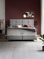 Ein luxuriöses Boxspringbett mit grauem Kopfteil und weißer Matratze in einem Schlafzimmer mit weinroter Wand.