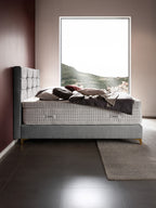 Ein Bett mit grauem Kopfteil und Matratze mit BettCo-Logo. Im Hintergrund ein Fenster mit Blick auf eine bergige Landschaft und Häuser.