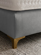Nahaufnahme eines grauen Boxspringbettes mit goldfarbenen Füßen und einer weißen Matratze auf einem Teppich.