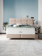 Ein beiges Boxspringbett mit einer weißen Matratze und grüner Bettwäsche in einem Schlafzimmer mit blauer Wand.