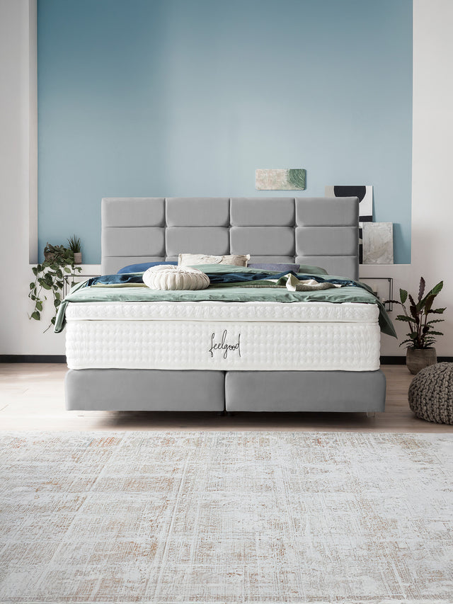 Ein modernes Schlafzimmer mit einem grauen Boxspringbett und einer weißen Matratze. Blauer Wandhintergrund.