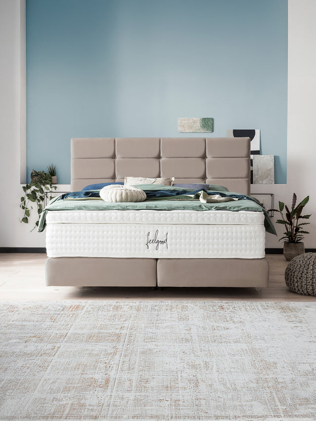 Ein modernes Schlafzimmer mit einem beigefarbenen Boxspringbett und blauer Wand. Auf dem Bett liegt eine weiße Matratze mit der Aufschrift feelgood.