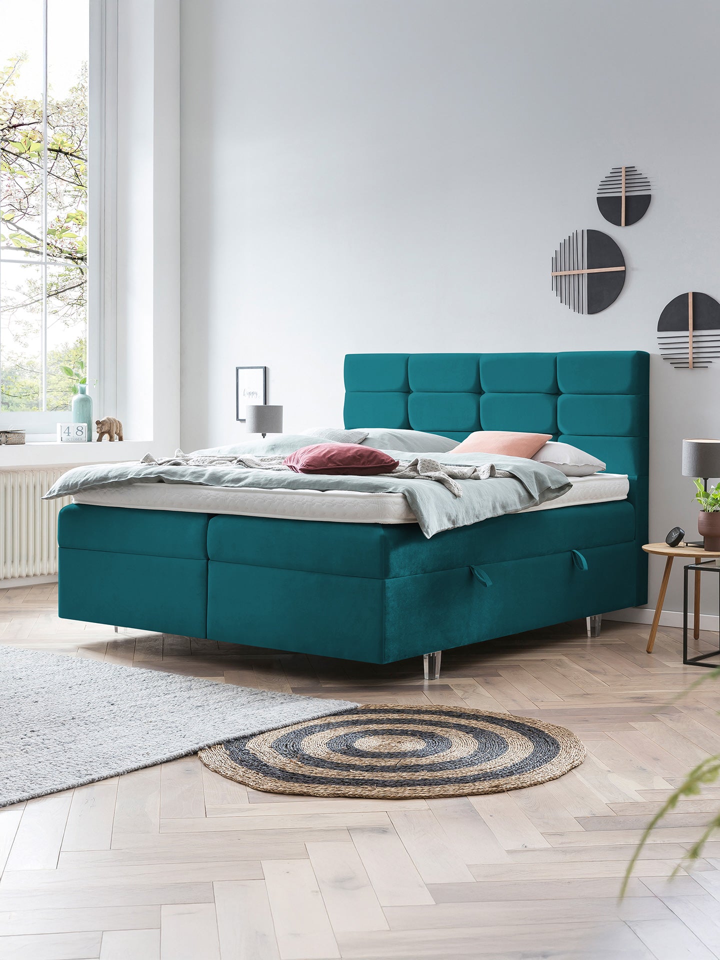 Türkises Boxspringbett mit gepolstertem Kopfteil in einem hellen Schlafzimmer mit Holzboden.
