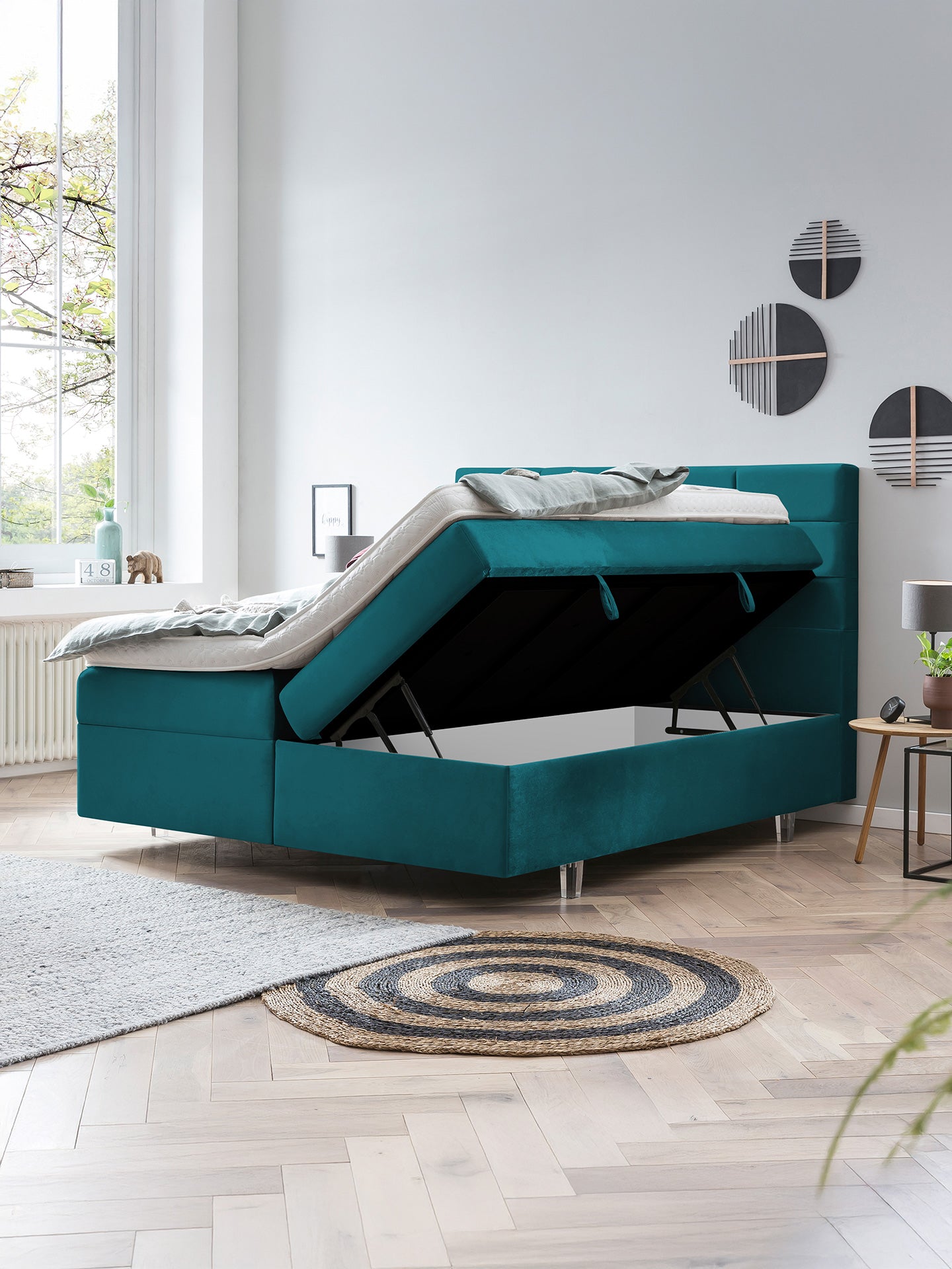 Türkises Boxspringbett mit offenem Stauraum und Bettzeug in einem hellen Schlafzimmer.