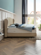 Ein Schlafzimmer mit einem Bett, das ein beiges Kopfteil und eine blaue Bettdecke hat. Das Zimmer hat Holzböden und ein großes Fenster.