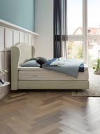 Ein helles Schlafzimmer mit einem BettCo Boxspringbett, blauer Bettwäsche und einem großen Fenster.