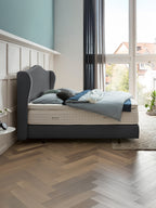 Ein Schlafzimmer mit einem Bett, das ein graues Kopfteil und eine blaue Bettdecke hat. Das Zimmer hat einen Holzboden und ein großes Fenster.