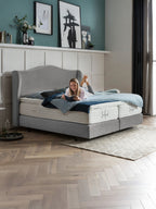 Eine lächelnde Frau liest ein Buch auf einem grauen Bett mit blauer Bettwäsche in einem modernen Schlafzimmer.