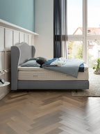 Ein modernes Schlafzimmer mit einem grauen Boxspringbett, blauer Bettwäsche und einem großen Fenster.