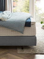 Ein Bett mit blauer Bettdecke und Überwurf, daneben ein Fenster mit Blick auf Bäume.