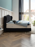 Schwarzes Bett mit blauer Bettwäsche und Kissen in einem Schlafzimmer mit Fischgrätparkett und großem Fenster.