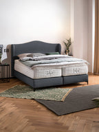 Ein Schlafzimmer mit einem grauen Boxspringbett, zwei Matratzen und einem grünen Teppich auf dem Holzboden.