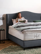 Eine lächelnde Frau liest ein Buch im Bett mit einem grauen Kopfteil und einer weißen Matratze.