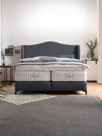 Ein modernes Schlafzimmer mit einem grauen Boxspringbett und weißen Matratzen. Auf dem Bett liegen Kissen und eine Decke.