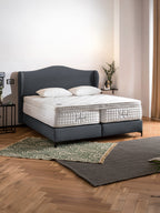 Ein Schlafzimmer mit einem grauen Boxspringbett und zwei weißen Matratzen. Auf dem Holzboden liegen zwei Teppiche.