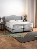 Ein graues Boxspringbett mit zwei weißen Matratzen und Kissen in einem Schlafzimmer mit Holzboden und Teppichen.