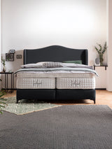 Ein elegantes Boxspringbett mit dunkelgrauem Kopfteil und weißen Matratzen in einem modernen Schlafzimmer.