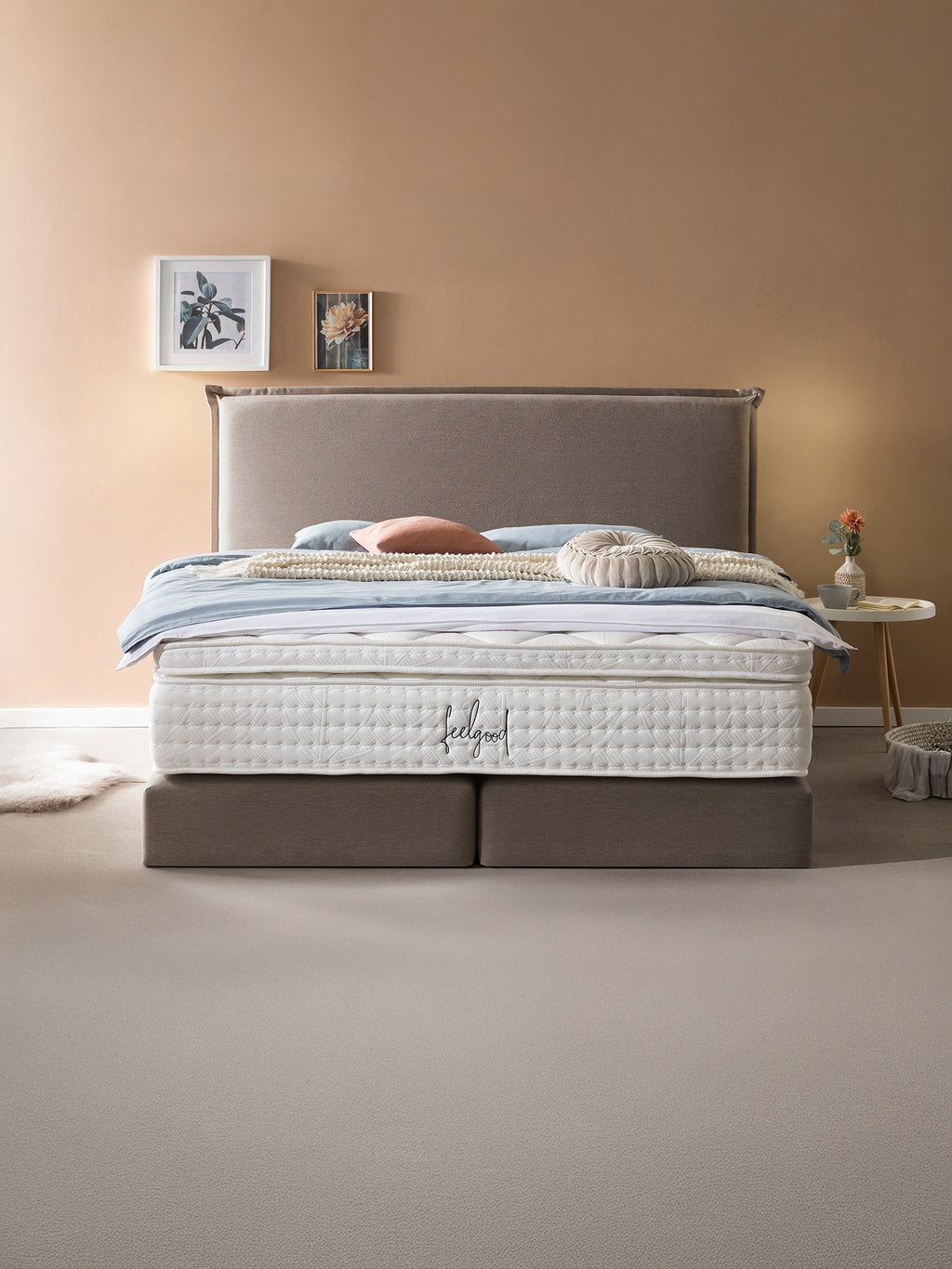 Ein luxuriöses Bett mit einem hohen, gepolsterten Kopfteil und einer dicken, weißen Matratze. Das Bett ist mit blauen und rosafarbenen Kissen sowie einer cremefarbenen Decke dekoriert. Daneben steht ein kleiner runder Tisch mit einer Vase und einer Tasse. Der Raum hat eine warme, beige Wandfarbe und einen hellen Teppichboden.