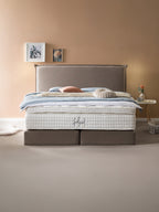 Ein luxuriöses Bett mit einem hohen, gepolsterten Kopfteil und einer dicken, weißen Matratze. Das Bett ist mit blauen und rosafarbenen Kissen sowie einer cremefarbenen Decke dekoriert. Daneben steht ein kleiner runder Tisch mit einer Vase und einer Tasse. Der Raum hat eine warme, beige Wandfarbe und einen hellen Teppichboden.