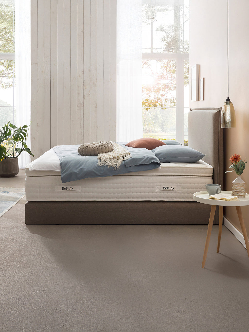 Ein Schlafzimmer mit einem Bett, das ein BettCo-Logo auf der Matratze hat. Das Bett ist mit blauen Kissen und einer Decke dekoriert.