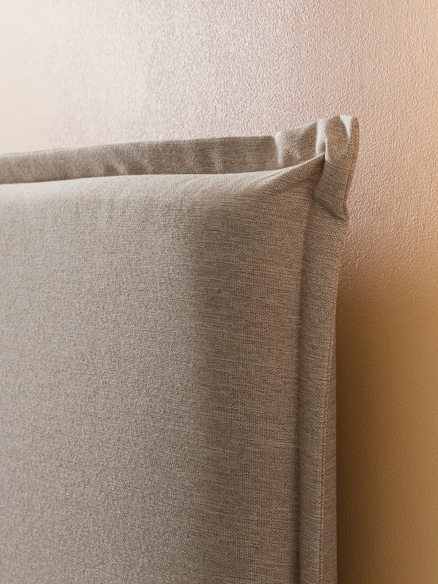 Nahaufnahme eines beigen Stoffes mit feiner Webstruktur, der als Kopfteil eines Bettes dient. Im Hintergrund ist eine hellbraune Wand zu sehen.