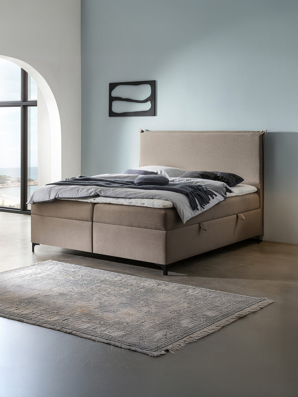 Ein Schlafzimmer mit einem beigefarbenen Boxspringbett, grauer Bettwäsche und einem Teppich auf dem Boden.