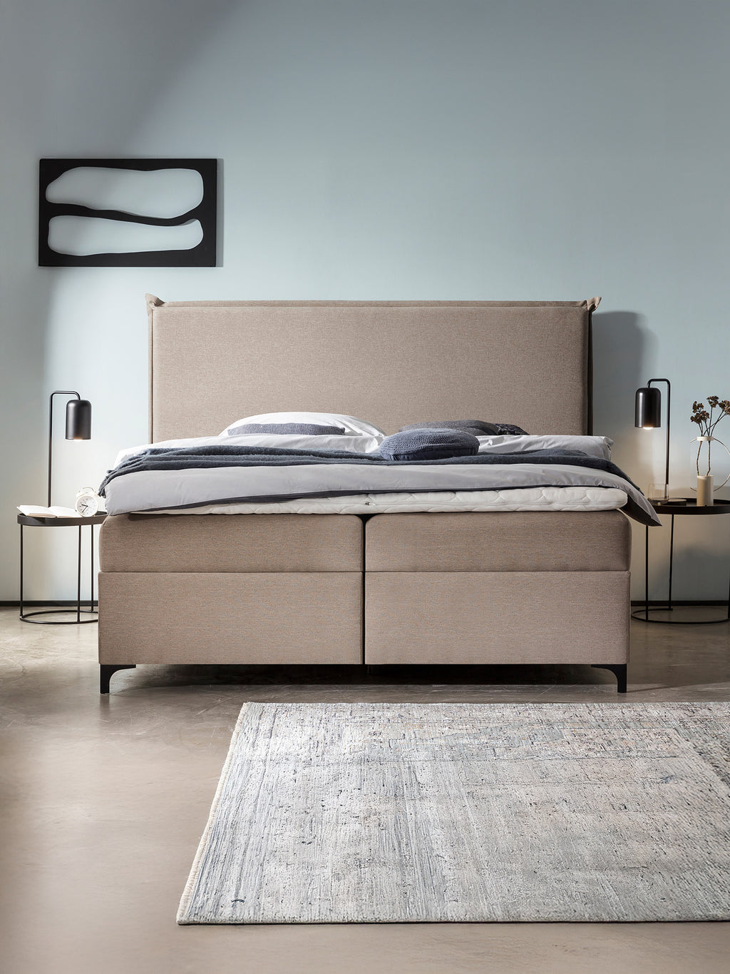 Ein Schlafzimmer mit einem beigefarbenen Boxspringbett, zwei Nachttischen mit Lampen und einem Teppich auf dem Boden.