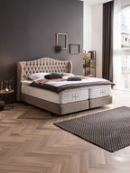 Ein Schlafzimmer mit einem großen, beigen Bett und einem dunklen Kopfteil. Ein Teppich liegt auf dem Holzboden.