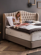 Junge Frau liest ein Buch im Bett mit einem gepolsterten Kopfteil und BettCo Matratzen.