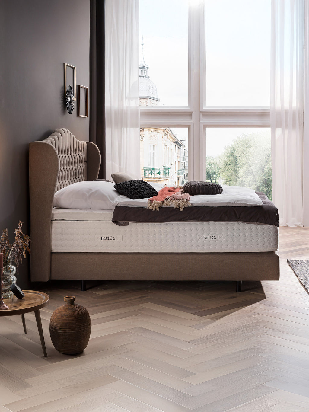 Ein Schlafzimmer mit einem Bett, das ein hohes, gepolstertes Kopfteil und eine BettCo-Matratze hat. Ein Fenster blickt auf eine Stadt.