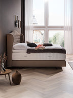 Ein Schlafzimmer mit einem Bett, das ein hohes, gepolstertes Kopfteil und eine BettCo-Matratze hat. Ein Fenster blickt auf eine Stadt.