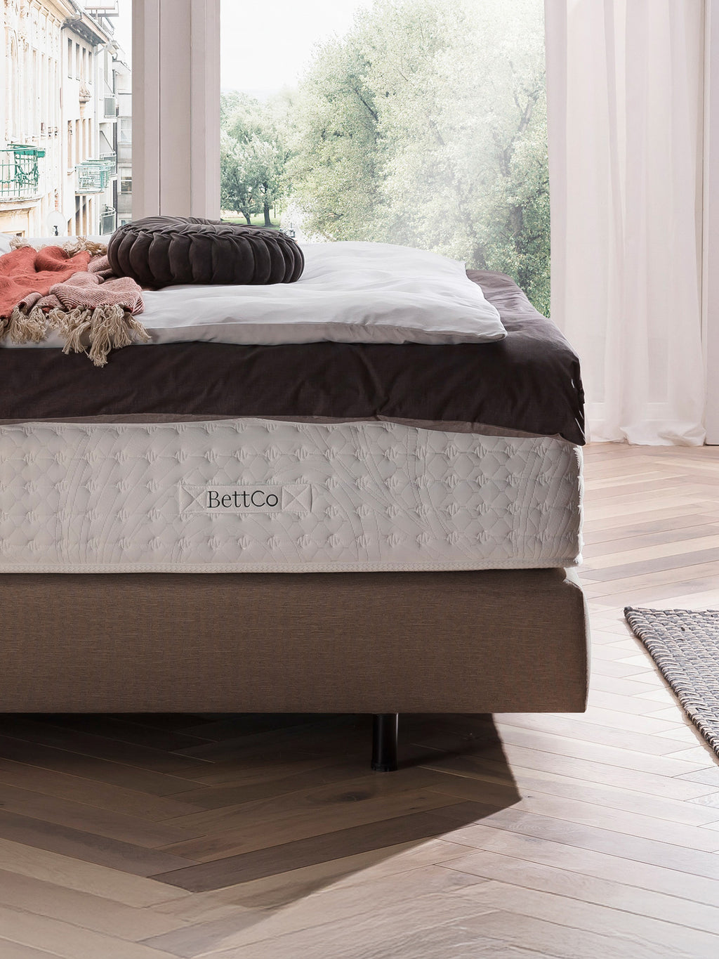 BettCo-Matratze auf einem Bettgestell mit Kissen und Decke, vor einem Fenster mit Blick auf Bäume.