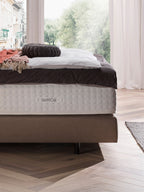 BettCo-Matratze auf einem Bettgestell mit Kissen und Decke, vor einem Fenster mit Blick auf Bäume.