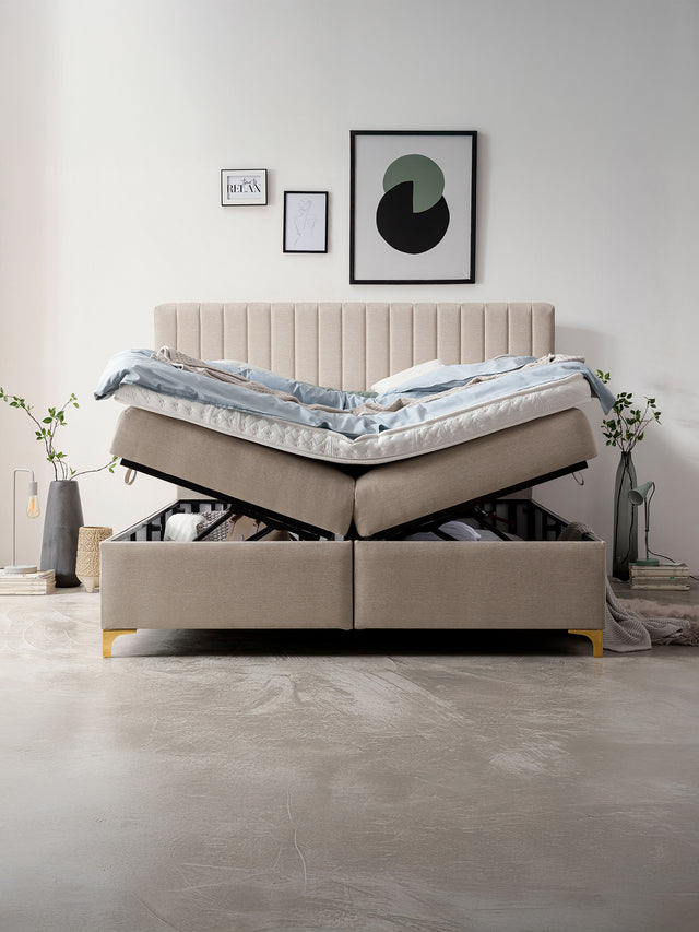 Beigefarbenes Boxspringbett mit offenem Stauraum, blauer Bettdecke und Kissen, umgeben von minimalistischer Dekoration.