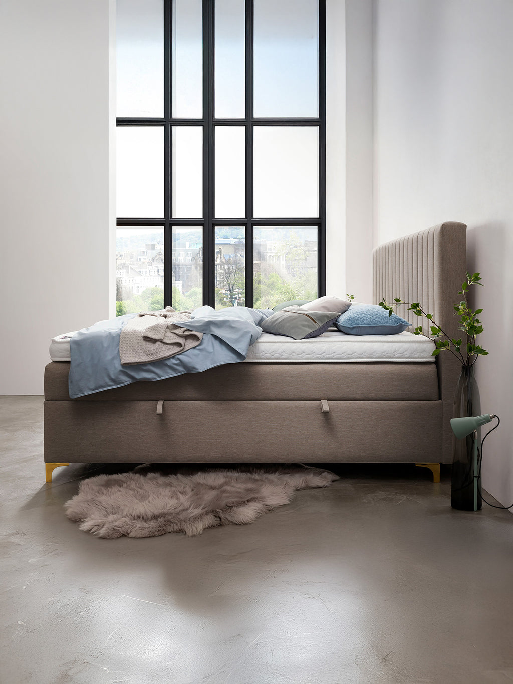 Ein Schlafzimmer mit einem großen Fenster, das einen Blick auf die Stadt bietet. Ein Bett mit Kissen und Decken steht im Raum.