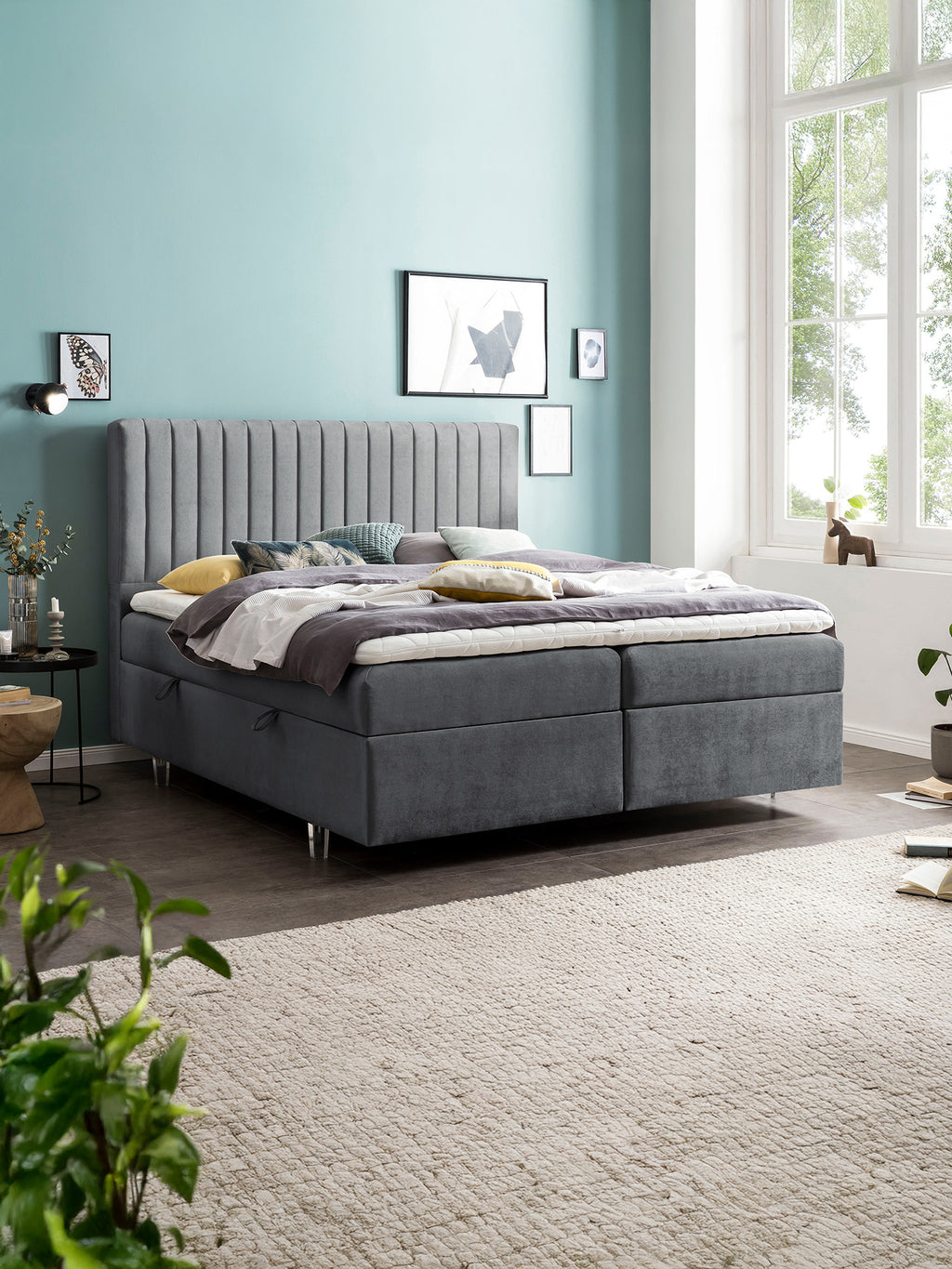 Modernes Schlafzimmer mit grauem Boxspringbett, hellblauer Wand und großem Fenster.