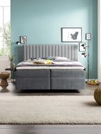 Graues Boxspringbett mit hohem Kopfteil in einem Schlafzimmer mit hellblauer Wand und Fenster.